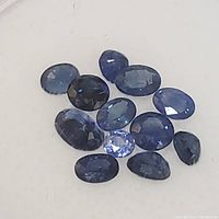 Photo showing 11 faceted sapphire gemstones in varying shades of blue and sizes, arranged on a white surface.