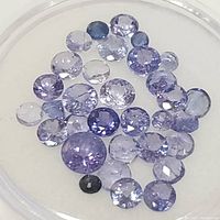 Top-down close-up of a round clear container holding 33 small faceted tanzanite gemstones of varying sizes and hues from pale violet to deep blue.