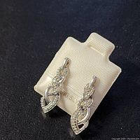 Photo of two silver earrings with intertwined loop design and diamond accent stones displayed on a white earring holder against black background.