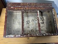 Top view showing front glass panels and wooden frame with painted lettering 'Simon's Havana Cigars'. Paint is chipped and some glass missing.