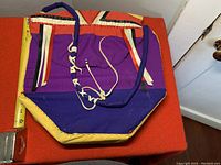 Custom handmade fabric ribbon bag showing purple, orange, and yellow colors with decorative hanging ribbons and a braided handle detail, measured against tape measure indicating approx 12 inches width.