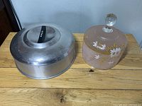 Photo showing the aluminum cake cover with black handle labeled 'CAKE' and pink frosted glass floral cake cover side by side on a wooden surface.