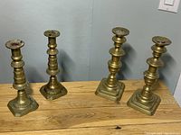 Four brass candle holders in varying designs placed on wood surface against plain wall.