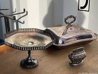 Silver plated octagonal pedestal server and covered rectangular serving dish on wooden table under natural light, showing surface tarnish and wear