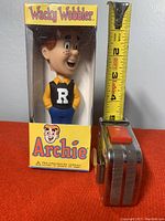 Front view of vintage Archie Wacky Wobbler bobblehead in original packaging next to a measuring tape showing approximately 6 inches tall.