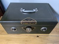 Front view of vintage dark green metal cash box/safe with combination dials and central lock, 'Superior Hand Safe' label visible.