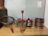 Photo showing vintage kitchen items with red handles on wooden surface including a flour sifter, nut chopper, hand mixer, wire mesh strainer, and cookie cutter.