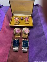 Closed box displaying two pairs of cufflinks with gold mesh bands and a single small round purple-pink button-style piece in the center.