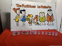 Vintage Flintstones advertising board with six character illustrations and French text with small pink collectible items arranged in front
