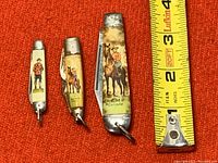 Three vintage RCMP folding knives with painted handles depicting mounted police and officers, displayed next to measuring tape showing sizes under 4 inches.