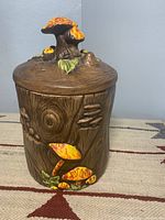 Side view of ceramic container showing wood grain texture with painted orange-yellow mushrooms attached and lid to top with cluster of mushrooms.
