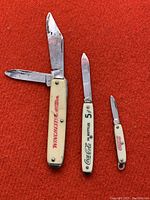 Three vintage advertising pocket knives on a red surface, showing the fronts with blades extended on two knives.