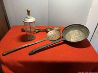 Wide view of vintage kitchen utensils on red cloth including a glass butter churn, ladles, scoops, and perforated spoon.