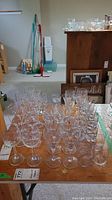 Photo shows multiple rows of clear glass stemmed glasses and tumblers arranged on a wooden table in a basement room with cleaning tools and furniture in background.