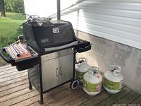 Front and side view of the Weber Spirit grill on cart showing cabinet, wheels, and three propane tanks beside it