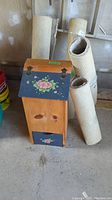 Wooden potato bin with floral design on lid and front, next to four rolled beige carpets standing vertically.