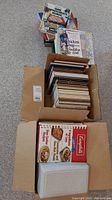 Photo showing two cardboard boxes filled with a large collection of cookbooks and recipe cards. Top boxes contain stacked cookbooks with various titles and colorful covers.