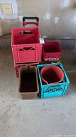 Photo showing red milk crate with an attached black-handled collapsible dolly, teal milk crate holding a red ceramic pot, brown rectangular crate, and several buckets including red and yellow Mil-Cre bucket.