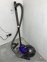 Full view of the Dyson DC37 vacuum cleaner upright with hose and brush head attached against a white wall and concrete floor.