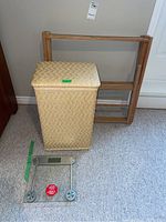 Photo showing woven clothes hamper, folding wooden clothes rack, and glass bathroom scale on carpeted floor.