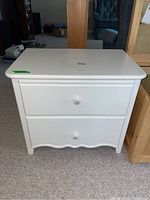 White night table with two drawers and floral-shaped drawer pulls, shown from the front and positioned on carpet.