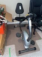 Front-left angled view of the silver and black recumbent exercise bike showing cushioned adjustable seat and pedals.