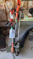 Wide photo showing three trimmers/edgers: two Black & Decker in orange/black and one gas-powered Weed Eater with a metal shaft, resting on the floor near tools and other garage items.