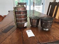 Photo showing the 9-inch tall antique copper tiffin carrier with multiple stacked compartments, latch, and handle, together with two smaller copper pots on a wooden table.