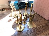 Lot of assorted brass items including two 10 inch tall candle sticks, several napkin rings, a brass mortar and pestle, and other brass pieces shown on hardwood floor with sunlight.