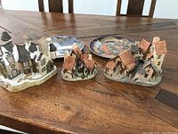 Three collectible village scenes by David Winter arranged on a wooden table along with two decorative plates, showing their scale and condition