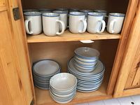 Shelf displaying seven off-white ceramic mugs with blue rims on upper shelf, below stacks of matching dishes and bowls.