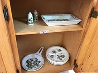 Wood cabinet shelf showing salt and pepper shakers, skillet-style dish, and six divided plates with a green and brown market design
