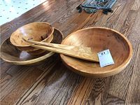 Full view of three wooden bowls and pair of wooden salad servers showing wood grain and size comparison on wooden floor.