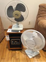 Overview of Dr Heater infrared space heater in wood-finish frame with front digital control panel, placed on floor along with two oscillating fans, one with black blades and one white, and remote plus manual on top.