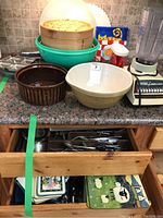 Photo showing counter with three nested stainless steel mixing bowls inside large green Rubbermaid mixing bowl with lid, bamboo steamer stacked on top, brown ceramic casserole dish, and cutlery drawers below containing utensils and kitchen tools. Colorful decorative plates in background.