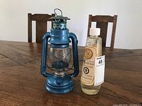 Blue metal oil lantern with glass globe and wire guard next to a plastic bottle of Heritage lantern oil on a wooden table