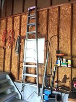 Full view of the aluminum ladder standing against a wooden-framed wall with tools and accessories nearby. The ladder has a pink top and 9 steps.