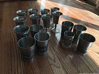 Full view of all 16 pewter drinking vessels arranged on wooden floor, showing variety in shapes and sizes.