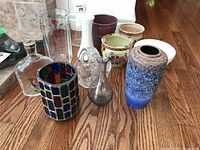 Photo of nine assorted vases and plant pots on wooden floor including a multicolored stained glass-style vase, hand-painted bottle, and crystal candy dish.