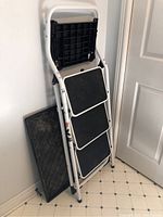 Folded white metal step ladder leaning against a wall, with the black boot mat behind it.