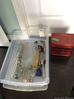 Plastic bin containing multiple necklaces including colorful beads, faux pearls, geometric and pendant designs, plus pairs of earrings, alongside a small red jewelry box with three drawers.