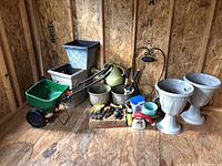 Full view of assorted garden items including two large pedestal urn planters, green broadcast spreader, watering can, gloves, hand tools, hose nozzle, leaf bags, and decorative wrought iron welcome sign/plant holder inside a wood-framed shed.