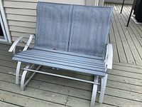 Front view of silver framed 2-seater outdoor glider bench on a wood deck, showing grey wooden slat seat and mesh cane backrest.