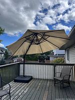 Large beige canvas umbrella open with black metal ribs and pole on wooden deck with two chairs nearby under blue sky and clouds.