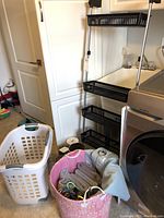Full view of laundry area showing over-the-door hanging rack with black plastic baskets, white laundry basket, and pink fabric basket with towels.