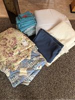 Photo of all linens including bedspreads, throws, duvet, and folded bed sheets on floor carpet.