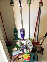 Photo showing multiple cleaning tools including Swiffer mops in green and purple, various cleaning cloths, brushes, and cleaning refills arranged on floor and leaning against wall.