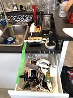 Photo showing kitchen sink, a drawer with assorted kitchen utensils, Magic Bullet blender components on counter, Nutrichef carving utensil in wooden holder, and Oster red electric pepper mill on counter.