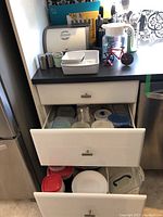 Overview of kitchen counter showing bread box, ceramic bakeware dishes, plastic storage containers in drawers, salt and pepper shakers, decorative pizza cutter, and utensils