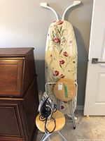 Photo showing the ironing board with floral pattern, Hamilton Beach iron, and two wooden folding stools stacked/stored together.